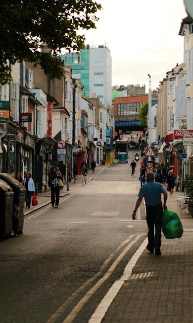 A busy urban street scene viewed from the sidewalk, with pedestrians walking along the pavement and crossing the road. In the foreground, a man wearing a blue shirt and dark trousers is seen carrying 