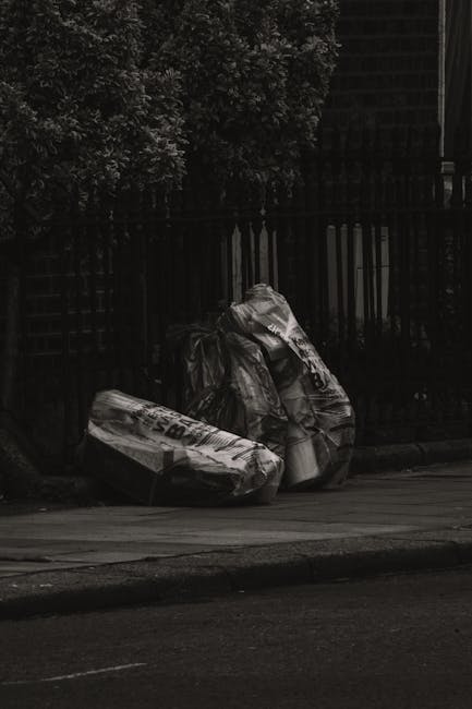 Two large black plastic garbage bags, filled and tied at the top, are placed on the pavement in front of a black metal fence with vertical bars and a brick wall behind it. The bags appear to contain v